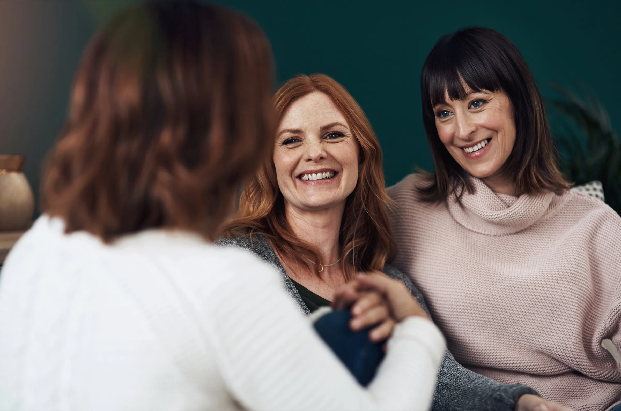 Two women smiling warmly at a friend as they sit together on a sofa